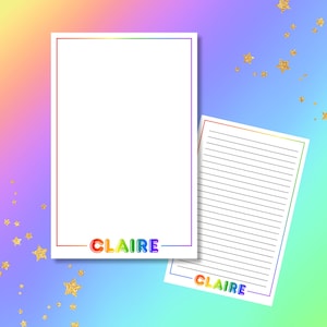 May include: Two white stationery sheets with rainbow borders and the name "CLAIRE" in colorful letters. One sheet is lined, the other is blank. The background is a gradient of rainbow colors with gold star accents.