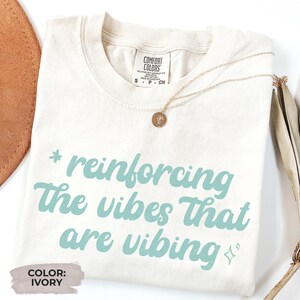 Puede incluir: Camiseta de color marfil con la frase "reinforcing the vibes that are vibing" en azul claro. Un collar dorado con un colgante está sobre la camiseta. La camiseta tiene la etiqueta "Comfort Colors".
