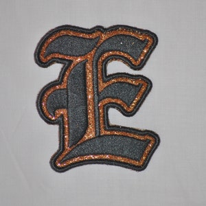 Op de afbeelding: Een strijkbare patch met de letter "E" in een gotisch lettertype, met zwart en gouden glitter.