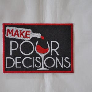May include: Black and red embroidered patch with white text that reads "Make Pour Decisions". The patch features a red wine glass with a red wine bottle pouring into it.