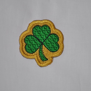 Op de afbeelding: Een geborduurde klaverblad-patch in groen met een gouden rand. De patch is perfect om kleding, tassen of andere accessoires een vleugje Ierse charme te geven.