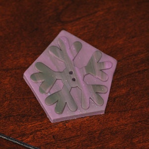 Puede incluir: Un botón en forma de copo de nieve de color morado y verde con una superficie texturizada.