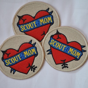 May include: Three embroidered patches with a red heart design and a blue banner that reads "SCOUT MOM" in yellow. The patches have a tan background and a circular shape, suitable for a scout mom.