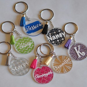 Könnte beinhalten: Sieben personalisierte Schlüsselanhänger mit verschiedenen Namen und Designs. Jeder Schlüsselanhänger besteht aus einem durchsichtigen Acrylkreis mit Glitzerhintergrund und einem weißen Namen oder Initial. Die Schlüsselanhänger sind an silbernen Schlüsselringen mit bunten Quasten befestigt.