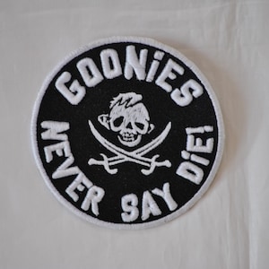 May include: Round black and white patch with the text "GOONIES NEVER SAY DIE!" in white lettering. The patch features a skull and crossbones design in the center, with a pirate theme.