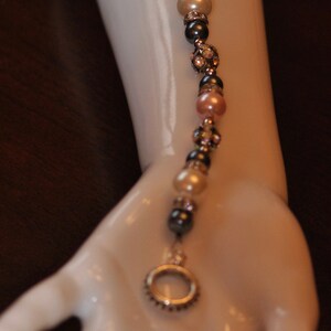 May include: A silver charm bracelet with black, white, and pink beads and a silver charm in the shape of a circle. The bracelet is on a white mannequin arm.