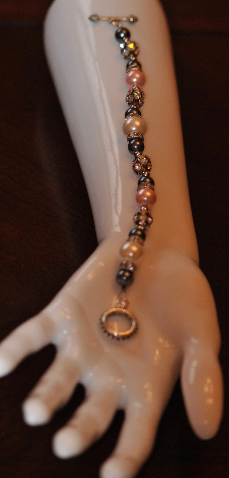 May include: A silver charm bracelet with black, white, and pink beads and a silver charm in the shape of a circle. The bracelet is on a white mannequin arm.
