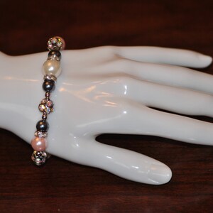 May include: A bracelet made of gray and silver beads with pink and white pearls. The bracelet is on a white mannequin hand.