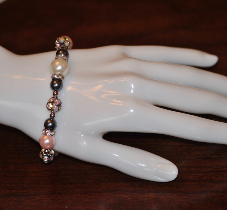 May include: A bracelet made of gray and silver beads with pink and white pearls. The bracelet is on a white mannequin hand.