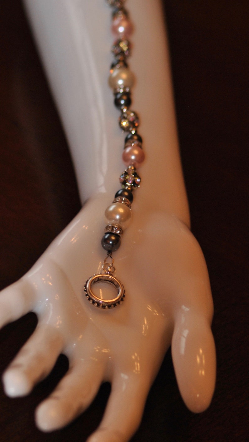 May include: A delicate bracelet with a silver ring charm, featuring alternating white pearls, pink beads, and dark gray beads.