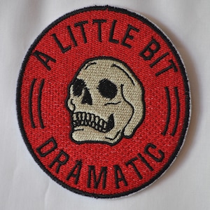 May include: An embroidered patch with a skull and the text "A Little Bit Dramatic". The patch is red and white.