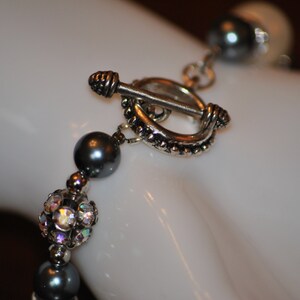 May include: A silver toggle clasp with a beaded design, attached to a gray pearl and crystal beaded bracelet.