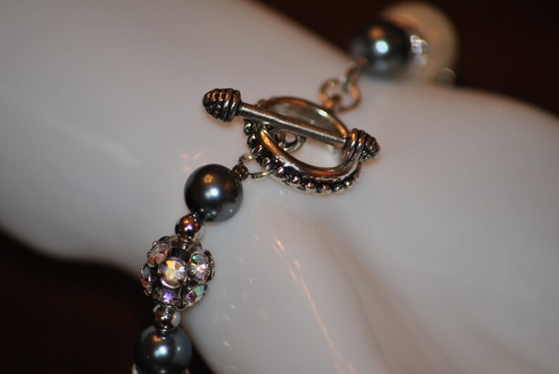 May include: A silver toggle clasp with a beaded design, attached to a gray pearl and crystal beaded bracelet.