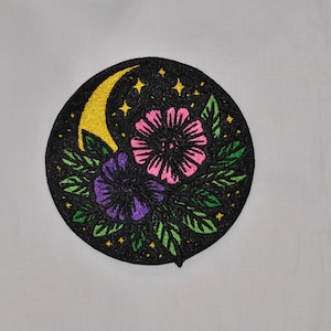 May include: Embroidered patch featuring a black background with a crescent moon and stars. Two flowers, one pink and one purple, are surrounded by green leaves. The patch is circular in shape.