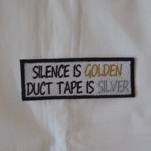 Op de afbeelding: Geborduurde patch met de tekst "SILENCE IS GOLDEN DUCT TAPE IS SILVER" in zwart, goud en zilver garen. De patch heeft een zwarte rand en is geplaatst tegen een witte achtergrond.