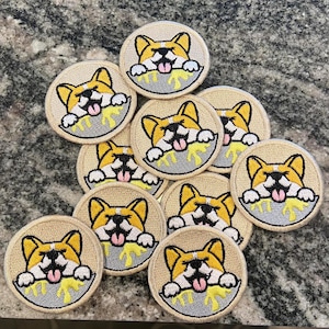 May include: A collection of ten embroidered patches featuring a cartoon corgi dog with its tongue sticking out, eating from a bowl of yellow food. The patches are beige with a grey outline and are approximately 5 cm in diameter.