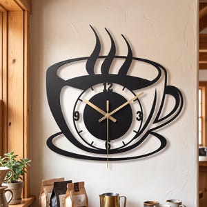 Puede incluir: Un reloj de pared negro con forma de taza de café con vapor saliendo de la parte superior. La esfera del reloj es circular y está centrada en el diseño de la taza, con manecillas finas de color dorado sobre un fondo negro.