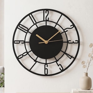 Puede incluir: Un reloj de pared redondo con marco de metal negro de diseño calado y grandes números negros estilizados. El centro es un círculo negro sólido con manecillas finas de color dorado para las horas y los minutos, creando un fuerte contraste.