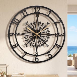 Puede incluir: Un gran reloj de pared circular de metal negro con un diseño central de rosa de los vientos con los puntos cardinales N, S, E, O, NO, NE, SO y SE. La esfera tiene números arábigos negros del 1 al 12 y manecillas de color dorado.