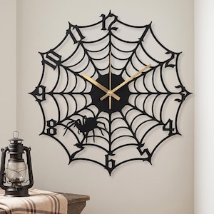 May include: A black wall clock shaped like a spider web with a central spider silhouette and gold-colored hands. The clock features large, gothic-style numerals integrated into the web design, creating a circular frame for the time display.