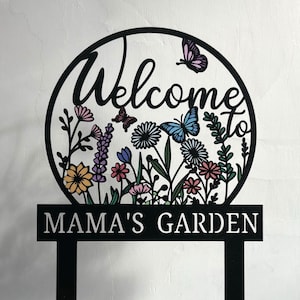 Puede incluir: Señal de jardín de metal negro con un diseño circular que presenta las palabras "Bienvenido al jardín de mamá" y flores y mariposas de colores.