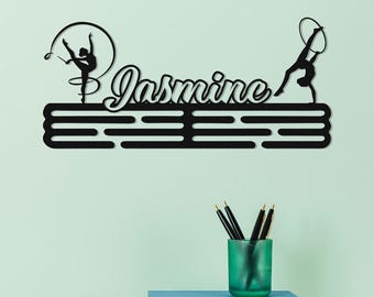 May include: Black metal wall art featuring the name "Jasmine" in cursive, with silhouettes of gymnasts performing with a ribbon and hoop. The design includes a horizontal bar for hanging medals or ribbons.