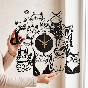May include: A black and white wall clock featuring a laser-cut design of multiple cats in various poses surrounding a central circular clock face. The clock face is white with black numerals and thin gold-colored hour, minute, and second hands.