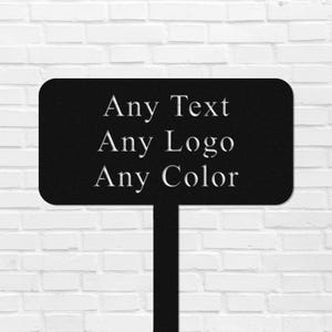 Op de afbeelding: Zwart metalen bord met de tekst "Any Text, Any Logo, Any Color" in het wit. Het bord heeft een rechthoekige vorm met afgeronde hoeken en een steel. De achtergrond is een witte bakstenen muur.
