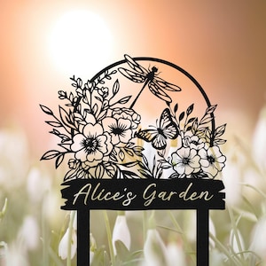 Puede incluir: Estaca de jardín de metal negro con un diseño floral, una libélula y una mariposa. La estaca tiene el texto "Alice's Garden". El diseño se encuentra sobre un fondo borroso de flores blancas.