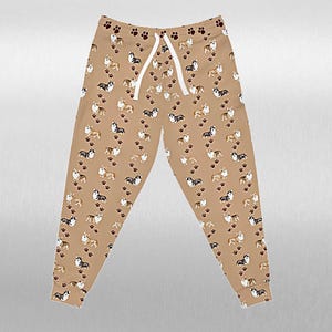 May include: Tan coloured sweatpants featuring a repeating pattern of corgi dogs and paw prints. The trousers have a drawstring waist and cuffed ankles. The corgis are in various colours, including brown, black, and white.