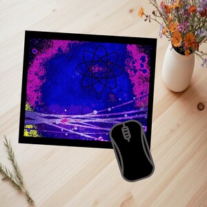 May include: A black computer mouse beside a rectangular mousepad with a black border. The mousepad features a blue and purple abstract design with an atom symbol in the centre. A small white vase with flowers is in the background.
