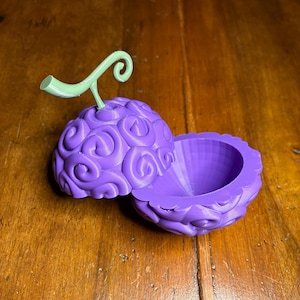 May include: A 3D-printed purple fruit with a green stem, split open to reveal a hollow interior. The fruit has a textured surface with a swirling pattern. The open fruit sits on a wooden surface.
