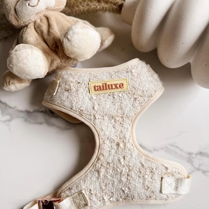 Könnte beinhalten: Ein beigefarbenes Hundegeschirr mit einer weißen, flauschigen Textur und einer goldfarbenen Schnalle. Das Geschirr hat ein gelbes Etikett mit dem Text "tailuxe" darauf.