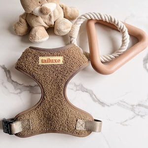 May include: A brown dog harness with the brand name "tailuxe" on a rectangular patch. A beige plush toy and a light brown triangle-shaped dog toy with a white rope are also in the image.
