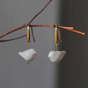 Op de afbeelding: Een paar gouden oorhangers met witte porseleinen vogelbedels.