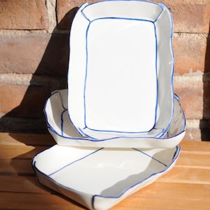 May include: Three white ceramic rectangular serving dishes with blue trim. The dishes are stacked on top of each other.