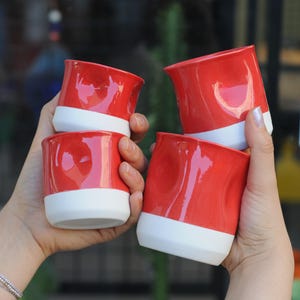May include: Four red ceramic mugs with white rims. The mugs have a textured, uneven surface and are stacked in a person's hands.
