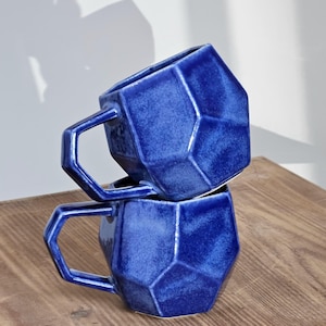 Op de afbeelding: Twee blauwe keramische mokken met geometrische ontwerpen. De mokken zijn op elkaar gestapeld. De mokken hebben een matte afwerking en zijn gemaakt van steengoed.