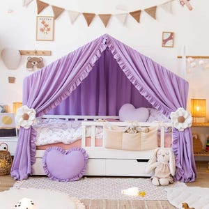 Puede incluir: Una cama infantil con dosel lila y elementos decorativos. La cama tiene un marco blanco, una almohada en forma de corazón y un conejito de peluche. La habitación está decorada con arte mural, una guirnalda y una alfombra.