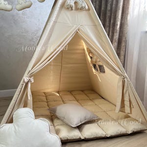 May include: A beige teepee tent with a matching floor mat and pillows. The tent has a small window and is decorated with cloud and rainbow-shaped ornaments. The text "Montes Wood" is visible on the tent.