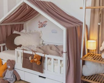 May include: A white wooden house bed with a canopy of mauve fabric. The bed is decorated with a moon-shaped pillow, stuffed animals, and a brown storage pocket. The wall features hot air balloon decals.