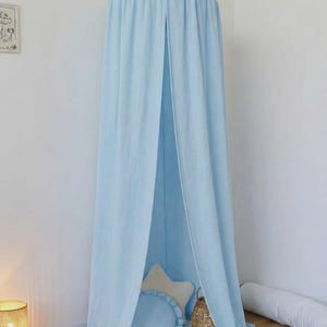 May include: A light blue fabric canopy suspended from the ceiling, featuring a conical top and long, flowing panels that drape down to the floor. The canopy creates a circular enclosure with a ruffled base, surrounding a small, round blue pillow.