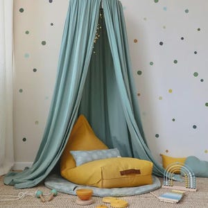 May include: A sage green fabric canopy hangs over a mustard yellow floor cushion and a round gray mat. The setup includes a light blue patterned rectangular pillow, a wooden rainbow abacus, a xylophone, and small wooden stacking toy pieces.