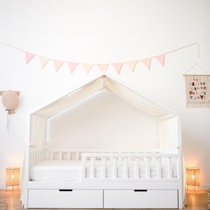 May include: A white canopy bed with two drawers, a white mattress, and a white headboard. The bed is decorated with a pink and cream-coloured banner. Two woven lamps flank the bed, and a leaf-shaped rug is on the floor.