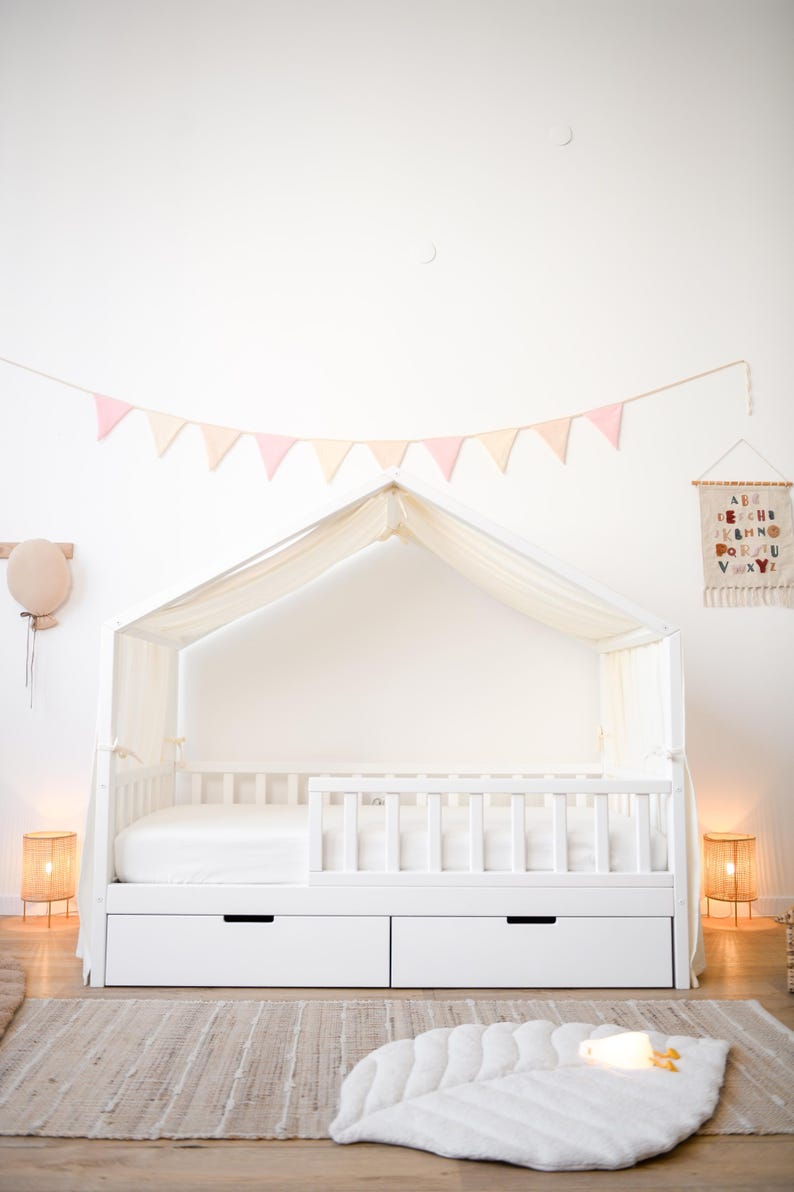 May include: A white canopy bed with two drawers, a white mattress, and a white headboard. The bed is decorated with a pink and cream-coloured banner. Two woven lamps flank the bed, and a leaf-shaped rug is on the floor.