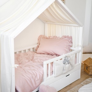 May include: A white wooden toddler bed with a canopy, featuring a pink duvet cover, pillow, and heart-shaped cushion. A white fabric pocket with a stuffed toy hangs on the side. The bed has two drawers and is on a wooden floor.
