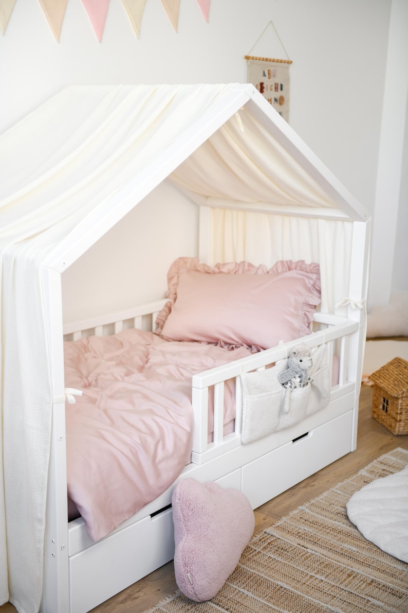 May include: A white wooden toddler bed with a canopy, featuring a pink duvet cover, pillow, and heart-shaped cushion. A white fabric pocket with a stuffed toy hangs on the side. The bed has two drawers and is on a wooden floor.