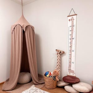 May include: A dusty rose fabric canopy hangs from the ceiling over a round woven rug and a textured floor cushion. Beside it, a tall giraffe-themed growth chart displays measurements in centimeters and feet, next to a basket of plush toys.
