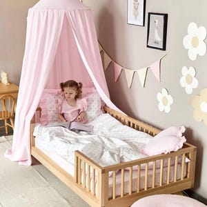 May include: A light pink fabric canopy hangs over a wooden toddler bed with slatted sides. The bed features white bedding with a small pink star pattern, a ruffled pink pillow, and a heart-shaped pink pillow. The canopy is suspended from the ceiling.