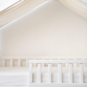 May include: A white wooden house-shaped bed frame with a white mattress and a white railing. The bed has two drawers at the bottom. A sheer, cream-coloured fabric drapes over the top of the frame. Pink and beige pennant flags are visible.